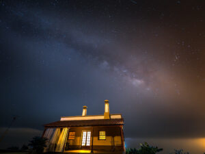 Night sky photograph of the Milky Way rising above a lit Canarian house, with a faint meteor streaking the right of the frame, La Palma.