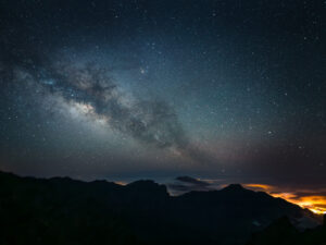 The Milky Way arching above silhouetted mountain ridges with distant town lights glowing through a sea of clouds, La Palma.
