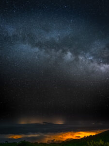 Vertical portrait of the Milky Way's galactic plane rising from the horizon over a sea of clouds lit by distant town lights, La Palma.