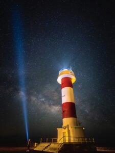 Night portrait of the Faro de Fuencaliente lighthouse beneath the Milky Way, with a figure shining a torch beam skyward.