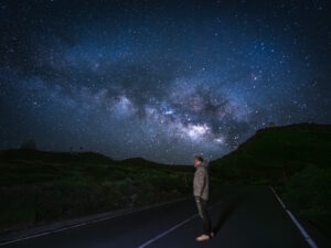 A figure standing on a mountain road at night, looking up at the Milky Way and distant observatory domes, La Palma.
