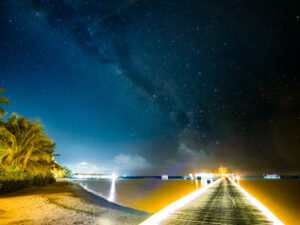 A lit wooden pier extending into the lagoon at night with the Milky Way rising above distant water villas, Maldives.