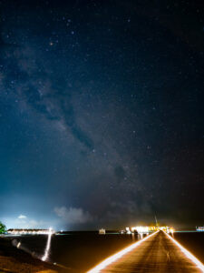 Long-exposure nightscape of a tropical beach pier with the Milky Way rising above water villas and distant clouds, Maldives.