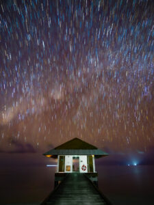 Long-exposure star trails curving above a wooden pavilion at the end of a pier in the Maldivian lagoon, Earth's rotation revealed.