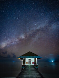The Milky Way rising vertically above a wooden pavilion at the end of a pier in a still Maldivian lagoon at night.