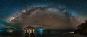 Panoramic photograph of the Milky Way arching over a thatched pier pavilion in a still Maldivian lagoon at night.