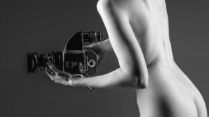 Black-and-white side-view nude study with the model holding a vintage medium-format camera at arm's length, sculptural curve of the back, plain studio backdrop.