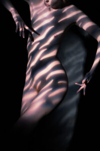 Colour fine-art nude with warm-toned horizontal stripes of light (as through Venetian blinds) drawn across the body, ethereal arms held outward, dark studio.