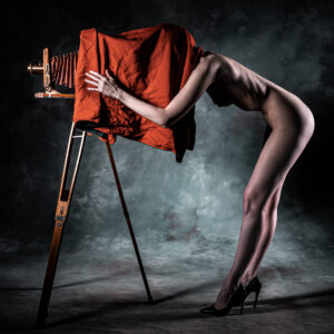 Colour fine-art nude in black heels bent forward with head under a red focusing cloth over a vintage view camera on a wooden tripod — a homage to photographic tradition.