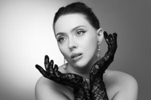 Black-and-white classic beauty portrait of a woman with hair pulled back, pearl drop earrings and black lace gloves framing her face, dramatic eye makeup.