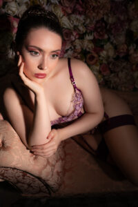 Colour boudoir portrait of a model in a floral tiara and plum lingerie reclining on a damask chaise with a wall of roses behind, romantic warm light.