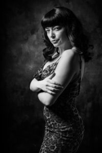 Black-and-white fashion-beauty portrait of Helen in a leopard-print slip dress, arms crossed, soft side light against a textured backdrop.