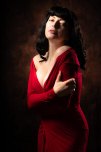 Colour fashion portrait of Helen in a red dress with one bare shoulder, arms wrapped across the body, dramatic chiaroscuro studio light.
