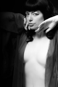 Black-and-white tasteful nude of Helen in a sheer open robe with hands raised by her face, sculptural lighting against a dark backdrop.