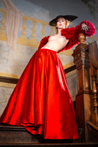 Colour avant-garde fashion portrait of Gemma on a grand staircase in a long red satin skirt, sheer red blouson sleeves, black hat and lace gloves.