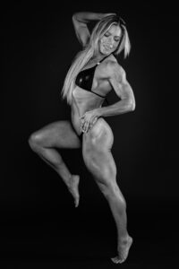 Black-and-white studio portrait of Lorna in a black bikini in an athletic dance pose, dramatic dark backdrop.