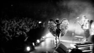 Black-and-white live music photograph: a performer in large feathered angel wings centre stage at a gig, band illuminated by stage lights to the right, crowd silhouetted at left.