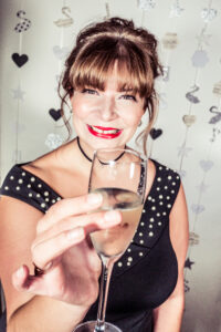 Colour photo-booth portrait at the Cygnet Players 20th anniversary celebration of a smiling woman in a black studded dress raising a champagne flute toward the camera.