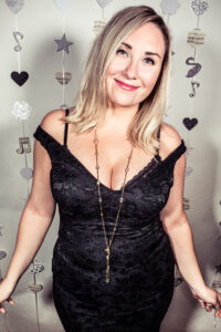 Colour photo-booth portrait at the Cygnet Players 20th anniversary celebration of a smiling blonde woman in a black sequined off-shoulder dress with a long gold pendant necklace.