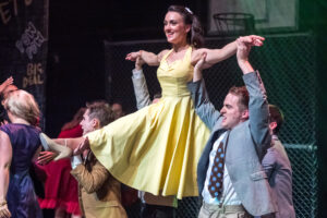 Theatrical performance photograph: a young woman in a yellow 1950s dress lifted by male dancers mid-routine in the Putney Light Opera Society (PLOS) production of West Side Story.