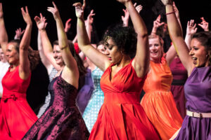 Theatrical performance photograph of the chorus of the PLOS production of West Side Story in jewel-toned dresses mid-routine with arms raised, central dancer the focal point.