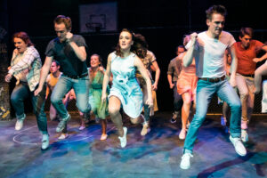 Theatrical performance photograph of the full company of the PLOS production of West Side Story dancing on the basketball-court stage set, 1950s costumes.