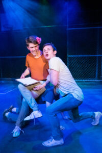 Theatrical performance photograph of two young male performers kneeling on the basketball-court stage of the PLOS production of West Side Story under blue light, one with a flower in his hair.