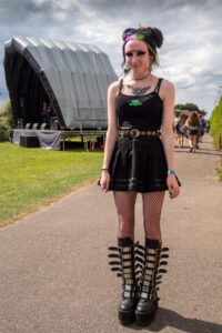 Colour portrait of a young woman in a black gothic dress, fishnet tights and platform boots posing in front of an inflatable festival stage, summer.