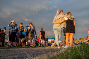 Festival photograph of two friends in shorts walking arm-in-arm into the Victorious Festival crowd at sunset, the giant "Victorious" letters in the distance, Portsmouth.