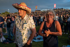 Festival crowd portrait at Victorious Festival, Portsmouth at sunset — a man in a sombrero and floral shirt next to a laughing woman in a vest, "Victorious" stage banners on the horizon.