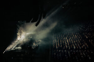 Black-and-white live music photograph from a high vantage point of Genesis on stage at a Manchester concert, shafts of stage light cutting through smoke onto the band, packed seated audience.