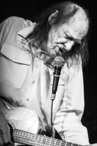 Black-and-white live music photograph of John Otway leaning into the microphone with an acoustic guitar, eyes closed, mid-vocal.