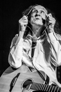 Black-and-white live music photograph of John Otway holding a curved wire above the microphone with both hands, eyes turned upward, acoustic guitar visible.