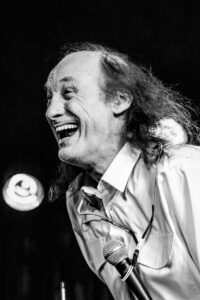 Black-and-white live music photograph of John Otway mid-laugh on stage, head thrown back, microphone in the foreground.