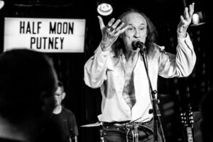 Black-and-white live music photograph of John Otway mid-incantation with hands raised, the "Half Moon Putney" sign behind him on stage.