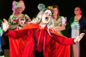 Theatrical performance photograph of the Queen Rat villain in red cape and mouse-ear crown, chorus in costume behind, from the PLOS production of Dick Whittington.