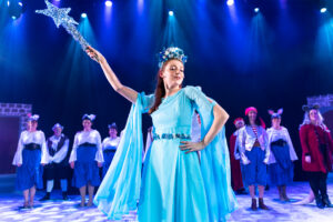 Theatrical performance photograph of a fairy in flowing blue robes and crown holding up a sparkling star wand, chorus arranged behind, from the PLOS production of Dick Whittington.