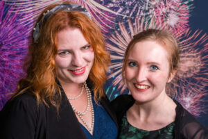 Photo-booth portrait of two smiling women — one with red hair in a black dress with pearl necklace, one in a dark green dress — at the Cygnet Players 25th anniversary, fireworks backdrop.