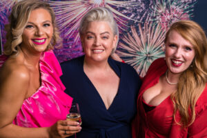 Photo-booth portrait of three smiling women in pink, navy and red dresses at the Cygnet Players 25th anniversary celebration, fireworks backdrop.