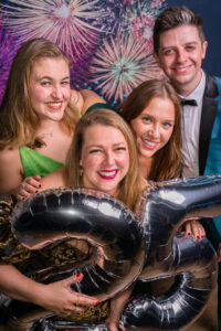 Photo-booth group photograph of four friends in colourful outfits behind giant silver foil "25" balloons at the Cygnet Players 25th anniversary, fireworks backdrop.