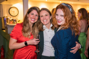 Colour candid photograph of three smiling women in red, black and blue dresses sharing a wine glass at the Cygnet Players 25th anniversary celebration.