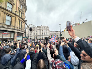 Colour photograph from within a crowd holding phones aloft in Trafalgar Square at the King's Coronation, May 2023, Union flags raised.