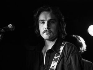 Black-and-white live music photograph of a young moustached frontman with a guitar strap at the microphone, the Troubadour, West Brompton, London.