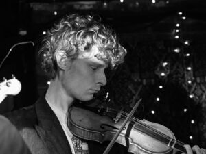 Black-and-white live music photograph of a curly-haired young violinist mid-play at the Troubadour, West Brompton, fairy lights behind.