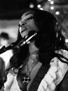 Black-and-white live music photograph of the same singer with eyes closed at the microphone at the Troubadour, West Brompton, statement coin-pendant necklace and ruffled blouse.