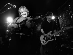 Black-and-white live music photograph of two young guitarists on stage at the Troubadour, West Brompton — one playing a white electric guitar, fairy lights behind.