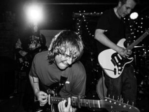 Black-and-white live music photograph of a young guitarist playing intently at the Troubadour, West Brompton, hair fallen forward, a second guitarist behind in stage lights.