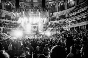 Black-and-white live music photograph from the back of the Royal Albert Hall stalls: the stage lit with "Damned" projected behind the band, audience filling the venue.