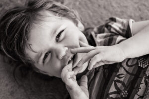 Black-and-white close-up of a young child lying down with a hand near the mouth, looking up at the camera with a playful expression.