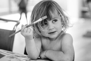 Black-and-white portrait of a young toddler with messy fair hair holding a paintbrush near her forehead.